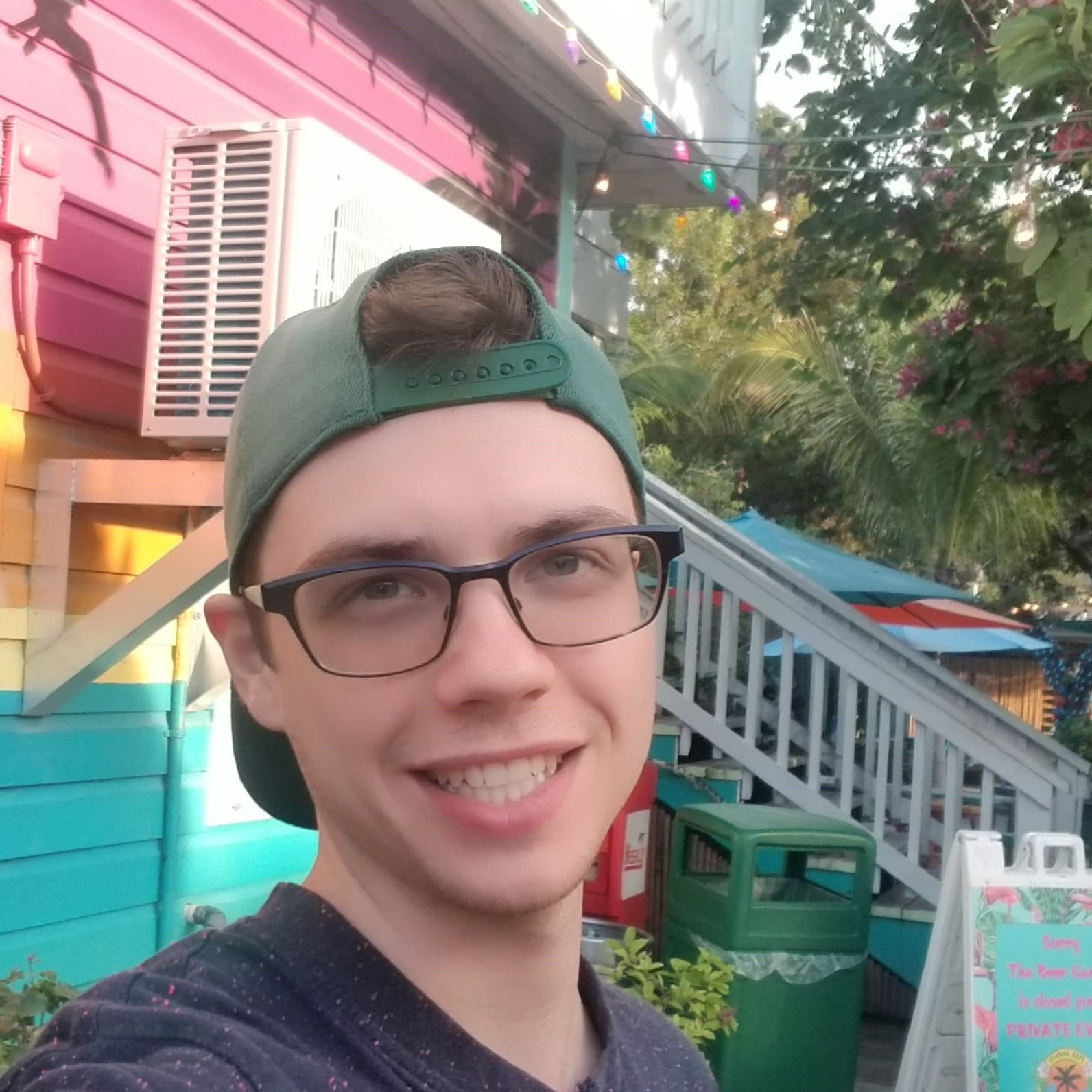 Paul with glasses and green hat smiling with colourful pink, yellow and cyan building close behind